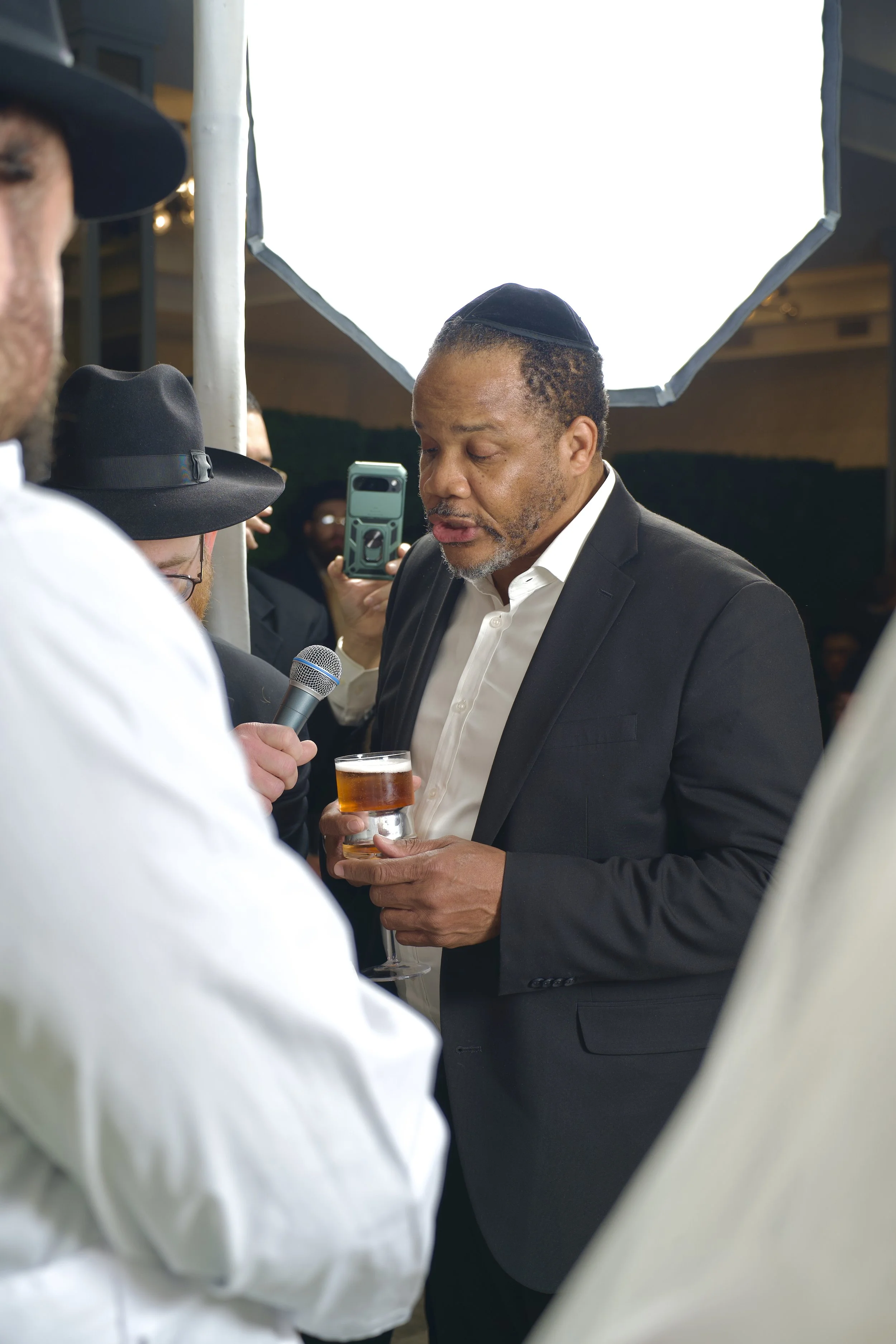 A man dressed in a black suit is being interviewed at a social gathering, holding a glass of beer, with a microphone held towards him by another person. There's a bright light diffusing through a large softbox overhead, and other guests are visible i
