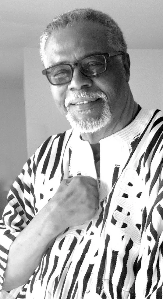 A smiling older man with glasses, a beard, and gray hair, wearing a patterned shirt, standing indoors.