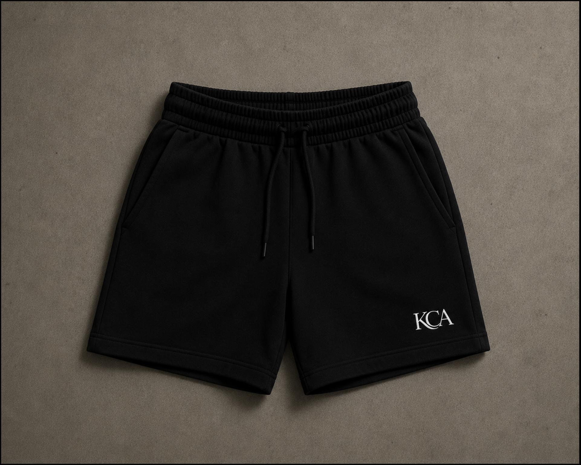 Black shorts with an elastic waistband, drawstring, and white monogram 'KCA' on the lower right leg, laid flat on a beige surface.