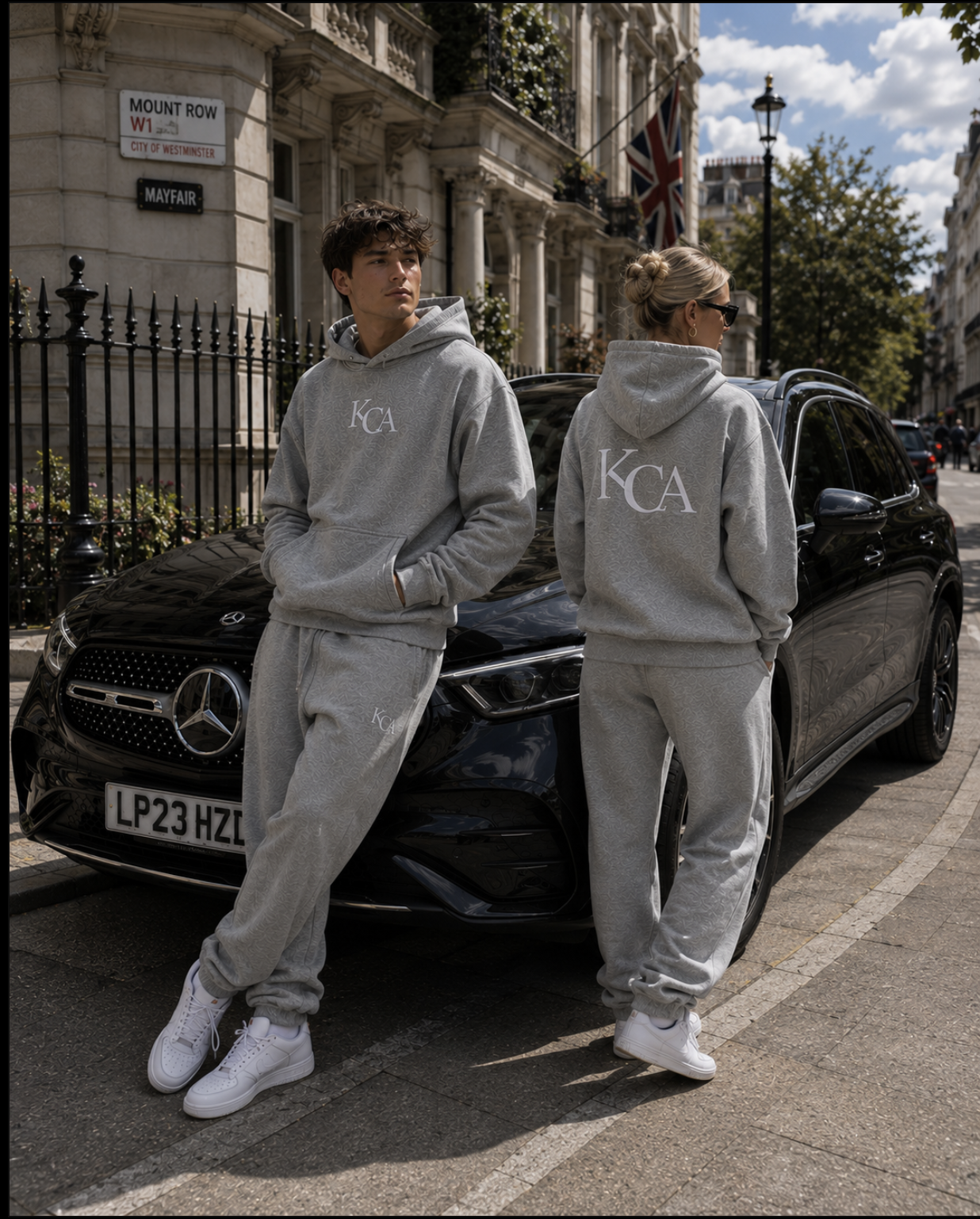 Two young adults, a man and a woman, are standing in front of a black Mercedes-Benz car on a city street with historic buildings. Both are dressed in matching light gray hoodies and sweatpants with the initials 'KCA,' and white sneakers. The man is leaning against the car with hands in his pockets, looking to the left, while the woman, with blonde hair in a bun and wearing sunglasses, has her hands in her hoodie pockets, facing away. There is a Union Jack flag hanging from a building, and a street sign indicating Mount Row in Westminster.