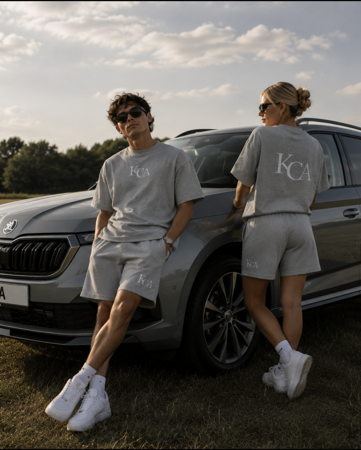 Two young adults, a male and a female, leaning against a gray car outdoors during the daytime. They are both wearing matching light gray outfits with 'KCA' logos, white sneakers, and sunglasses. The male has brown curly hair, hands in pockets, and is slightly leaning back, while the female has blonde hair tied in a bun and is facing sideways with her arms crossed.