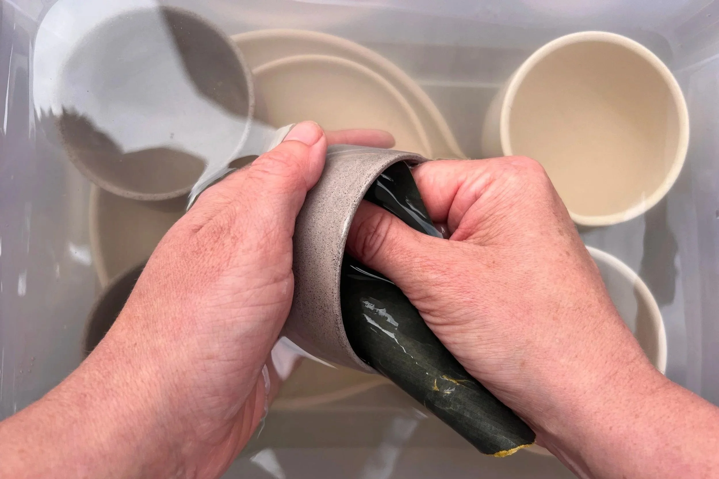 Person holding and pouring the contents of a ceramic mug into a sink with other mugs piled in the background.
