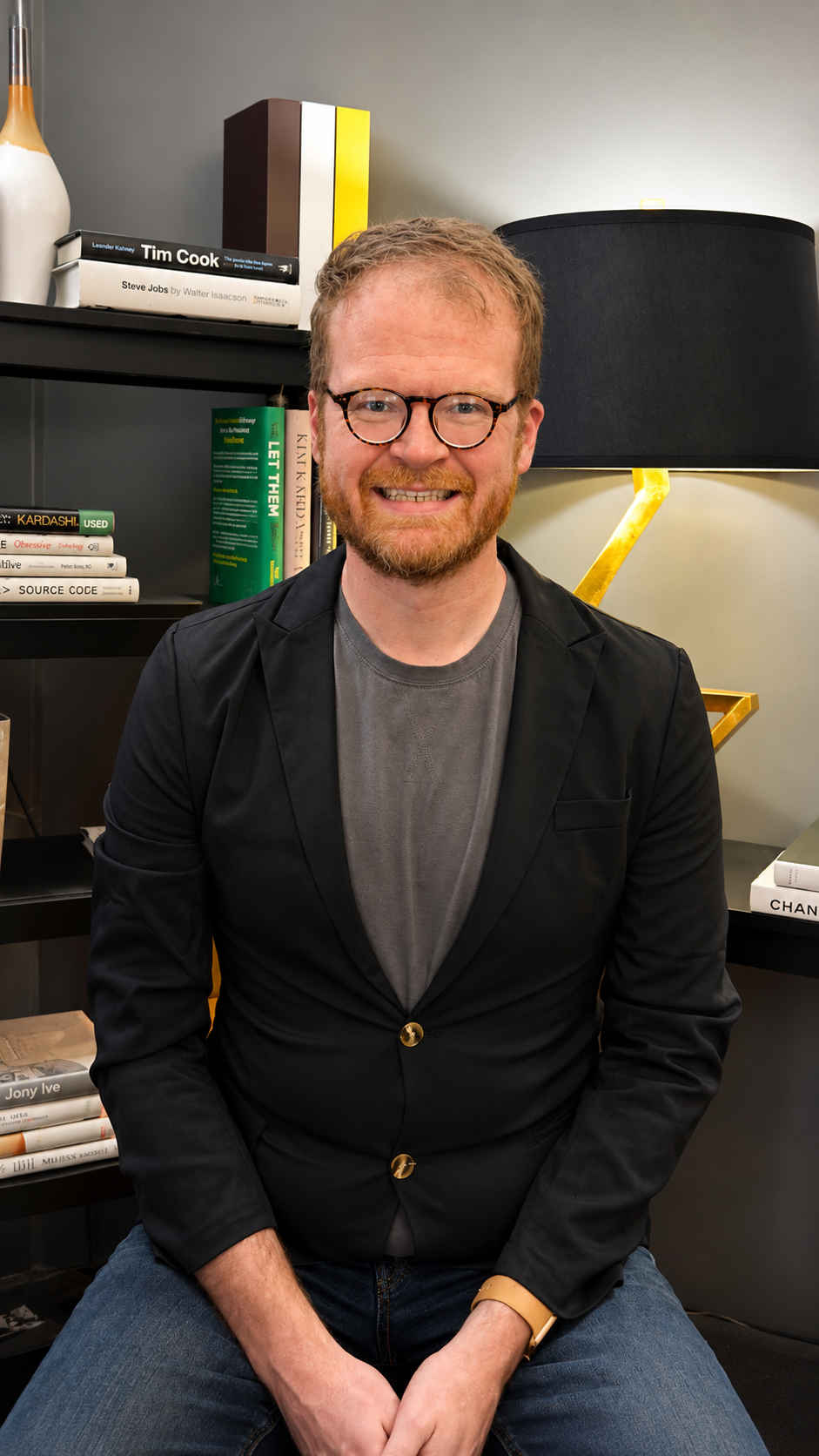 A smiling man with glasses, a beard, and curly hair, wearing a black blazer and gray t-shirt, sitting in front of a bookshelf with books and decorative items, in a modern room with a black lamp and a gold accent.