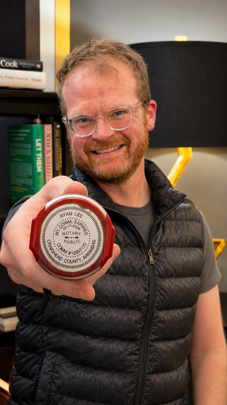 Smiling man with glasses and a beard holding a notary seal with his name on it, indoors with books and a black lamp in the background.