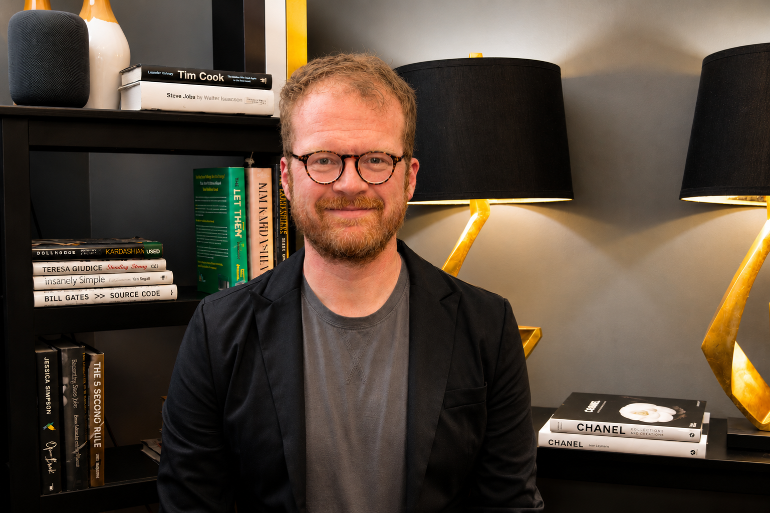 A man with glasses and a beard smiling indoors. Behind him is a black bookshelf with books and decorative items, and two black lamps with gold stands on the wall.