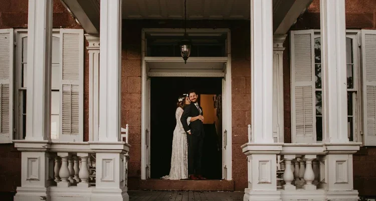 A couple standing on a porch in front of a house, with the woman hugging the man from behind.