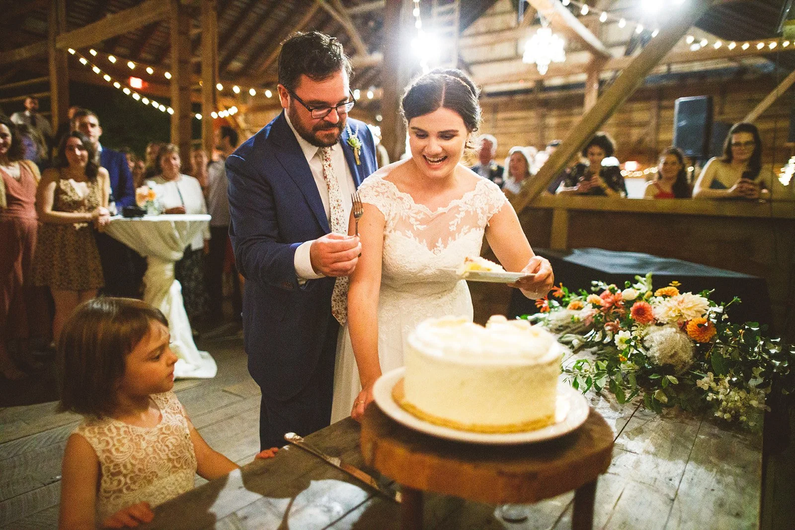 Bride and groom cutting wedding cake, surrounded by guests in a rustic barn setting decorated with string lights and flowers.