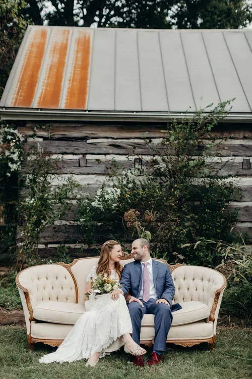 A bride and groom sitting on a vintage cream-colored sofa outdoors, smiling and looking at each other, with greenery and a rustic wooden barn background.