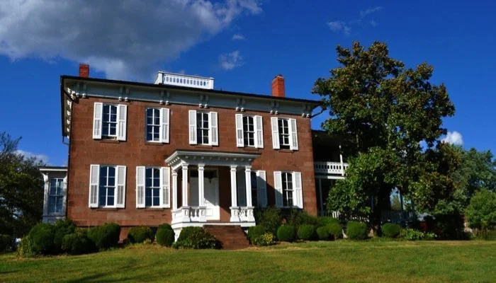 A large historic brick house with white shutters, a porch with columns, a staircase, and a neatly maintained lawn with bushes and trees under a partly cloudy sky.