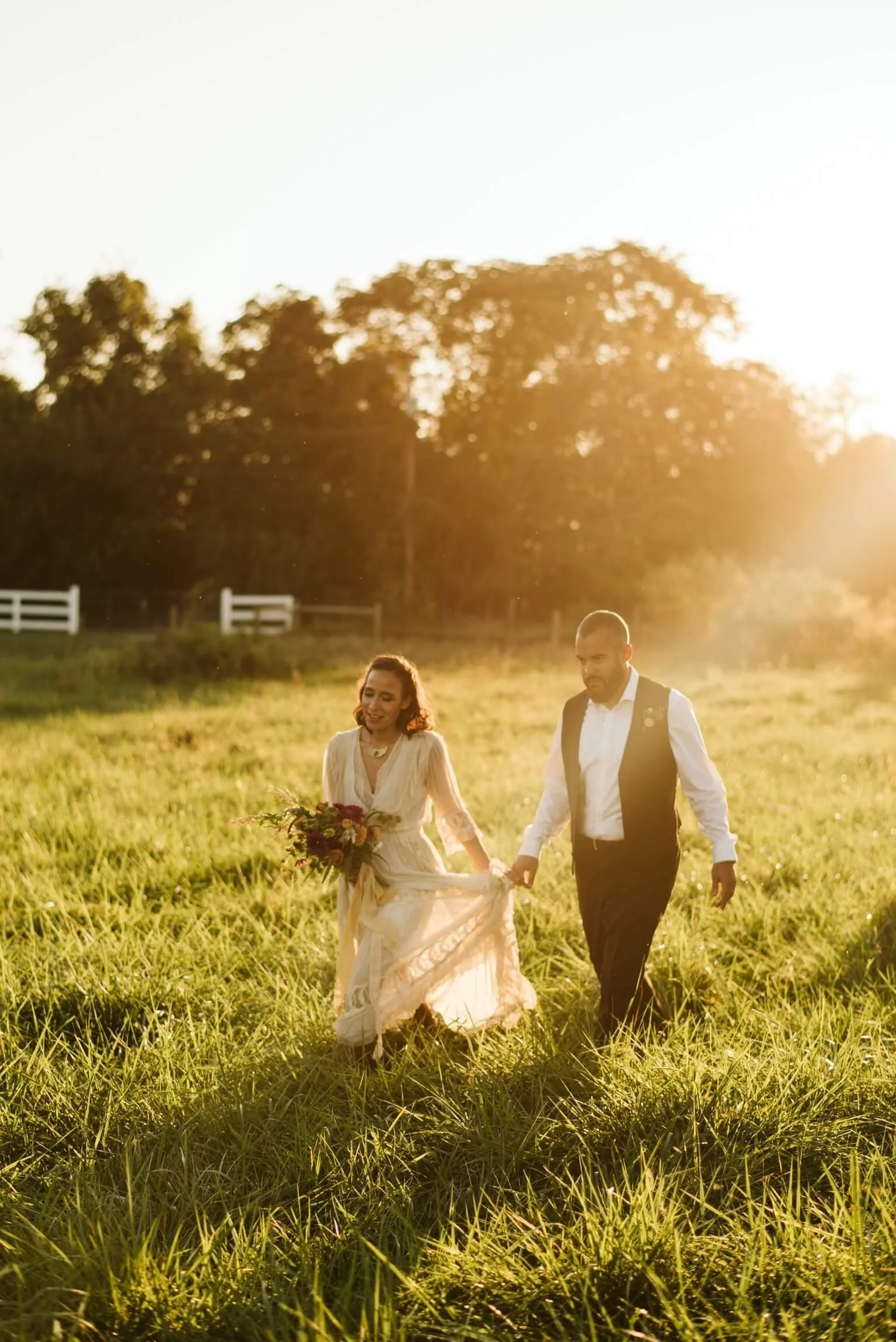 A bride and groom walk hand in hand through a grassy field during sunset  at Rocklands Farm Winery, dressed in wedding attire, with trees in the background and sunlight filtering through.