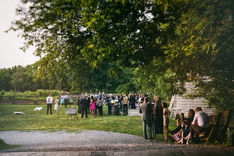 Outdoor gathering of people under trees, socializing near a white wooden building, with some sitting on benches and others standing in groups.