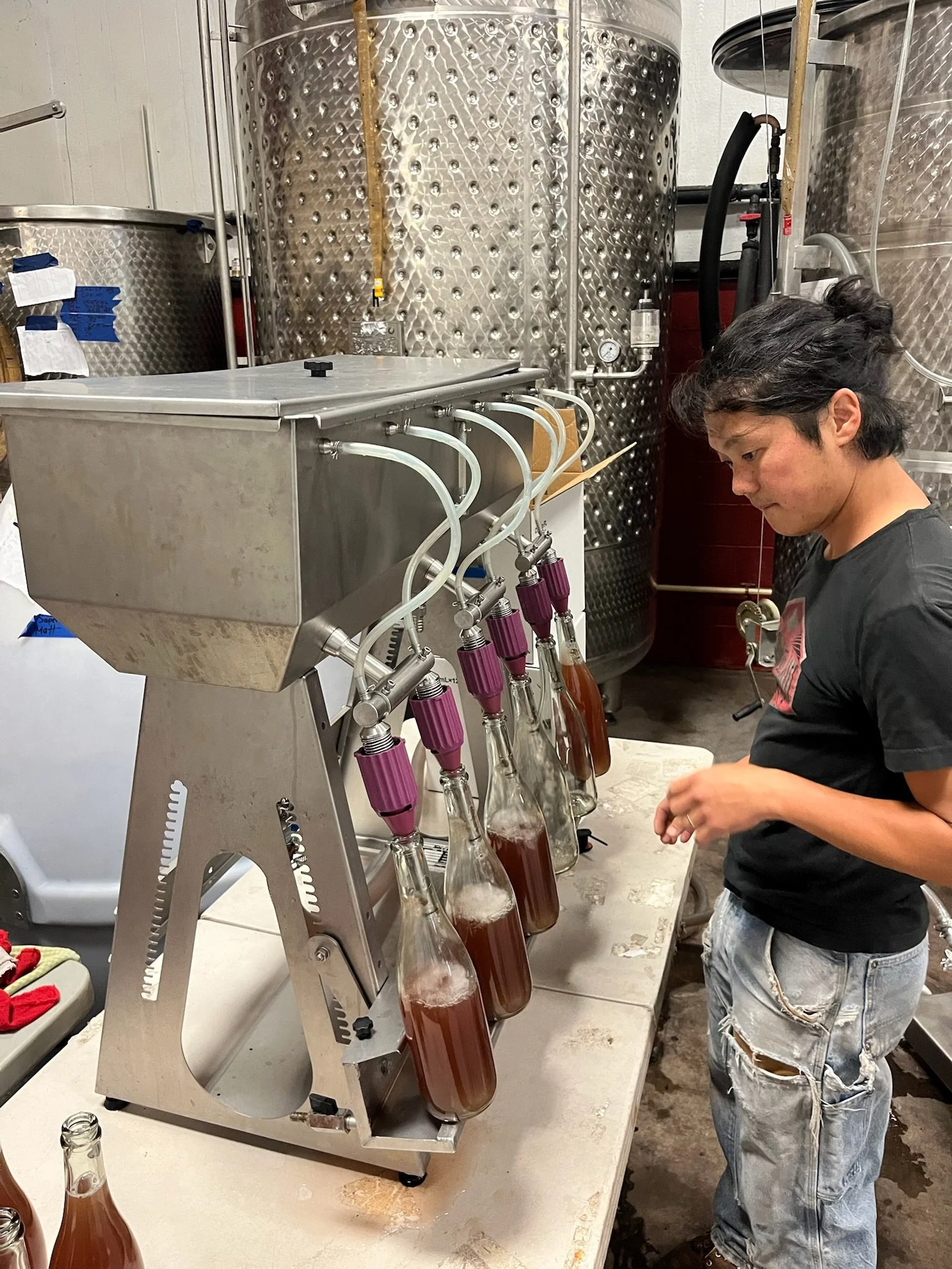A person making kombucha using a continuous brew system at a brewery or distillery. They are pouring or handling bottles filled with a dark amber liquid, likely kombucha.