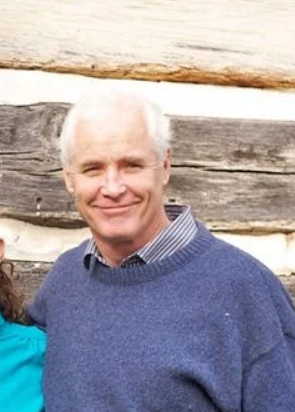 A photo of an older man with white hair, smiling, wearing a blue sweater and a collared shirt, standing outdoors in front of logs.