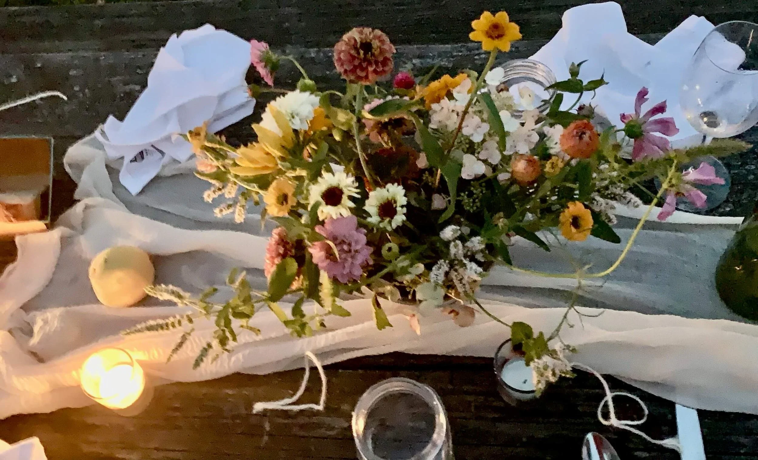 A floral arrangement with various small flowers in a glass vase, surrounded by candles and decorative objects on a table, with a dark background.