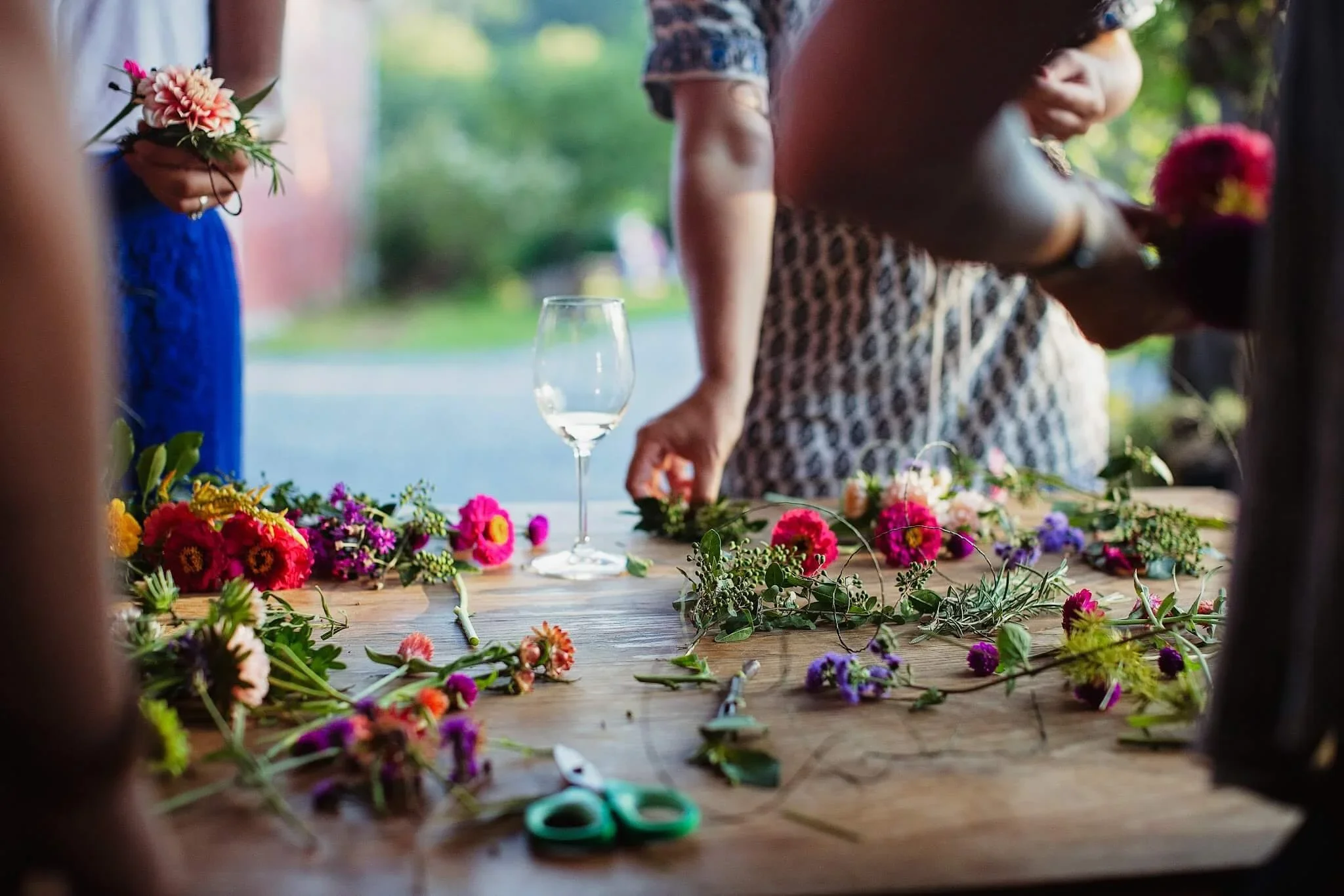 People preparing floral arrangements on a wooden table outdoors, with various colorful flowers, scissors, and a wine glass, in a garden or park setting.