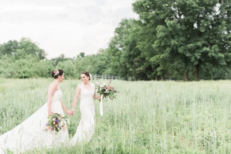 Two women in white wedding dresses holding bouquets, walking hand in hand through a grassy field with trees and a small bridge in the background.