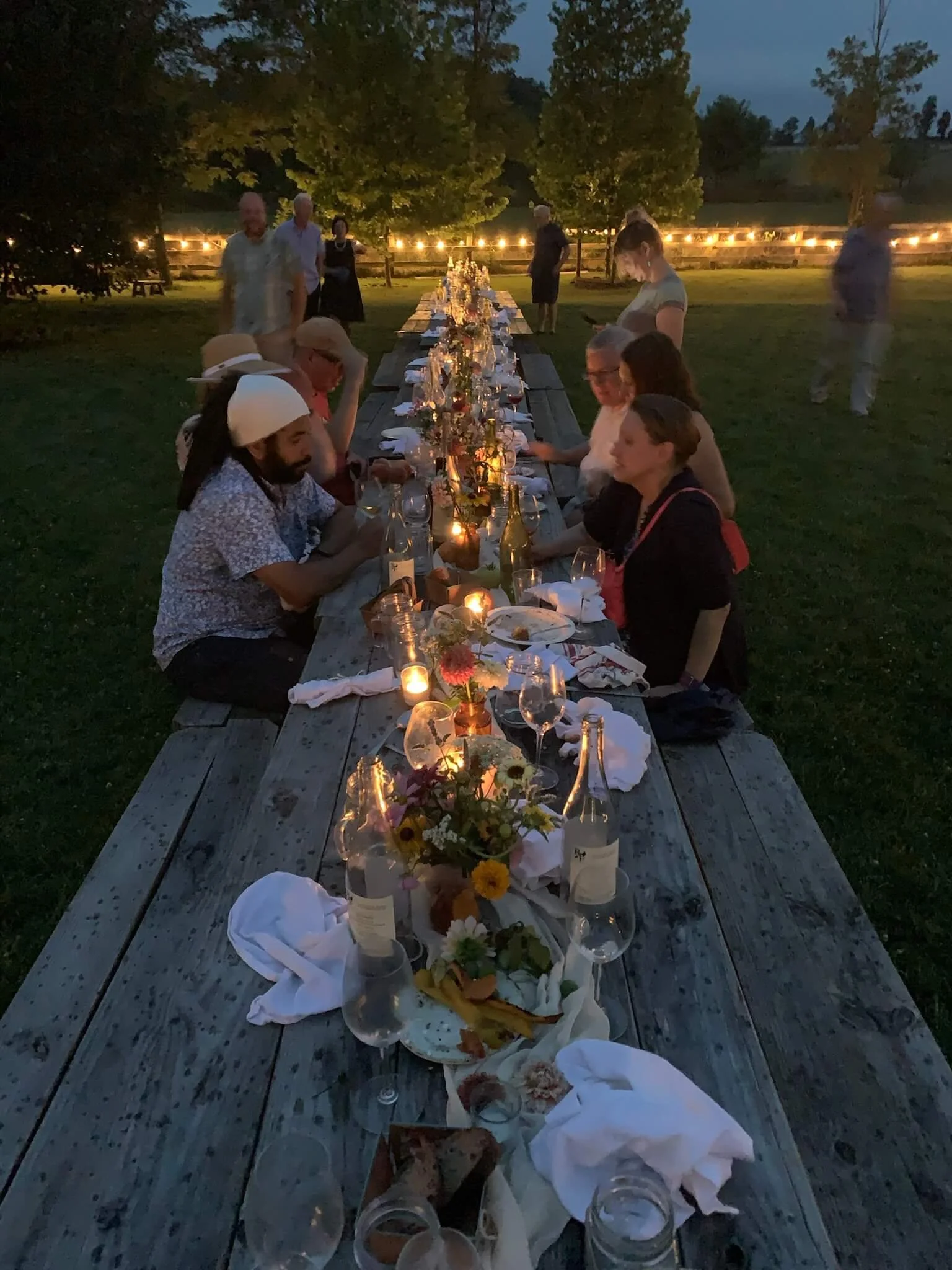 A long outdoor dining table decorated with flowers, candles, and wine glasses at dusk, with people seated and standing around, in a grassy area with trees and string lights in the background.