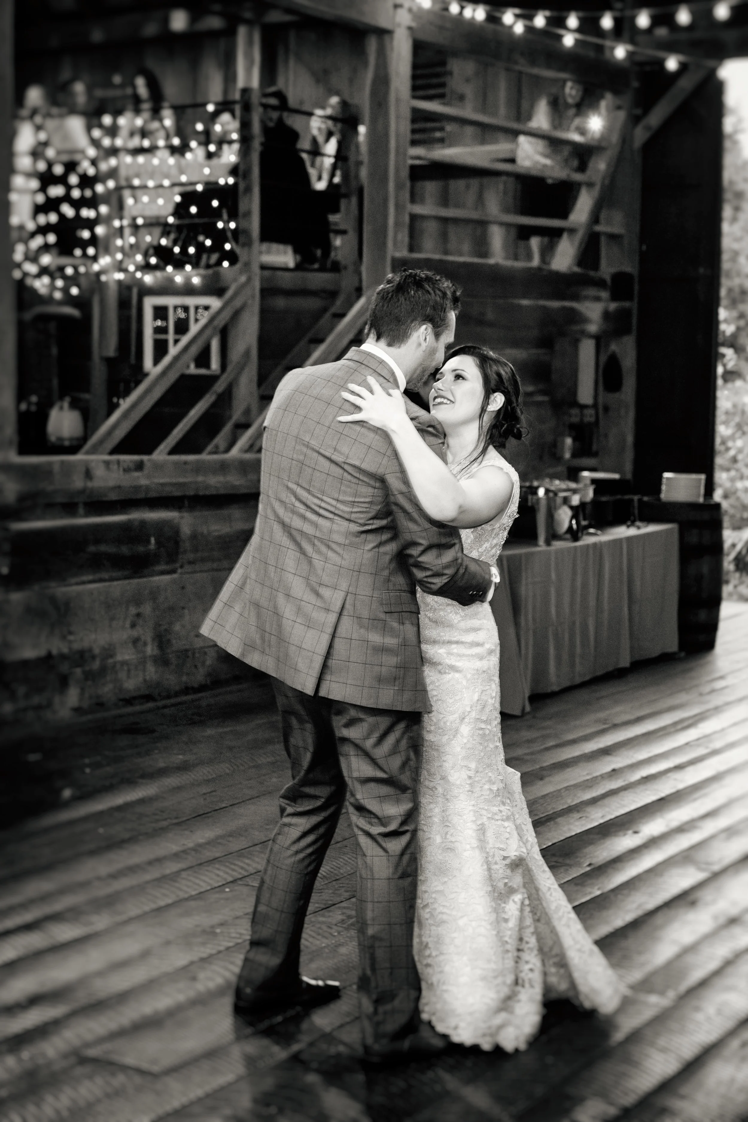 A black and white photo of a couple dancing closely, with the woman in a lace wedding dress and the man in a suit, on a wooden floor with string lights overhead, in a rustic venue with wooden structures and a staircase in the background.