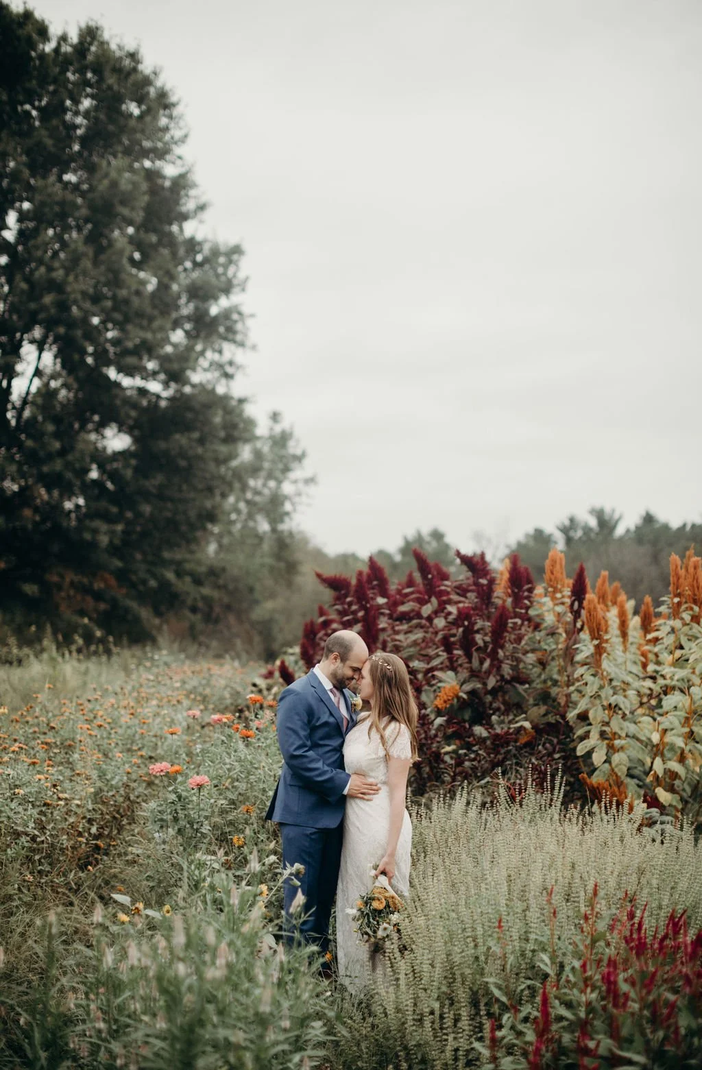 A bride and groom standing close together in a garden full of colorful flowers and plants, with trees in the background, during a wedding or romantic photoshoot.