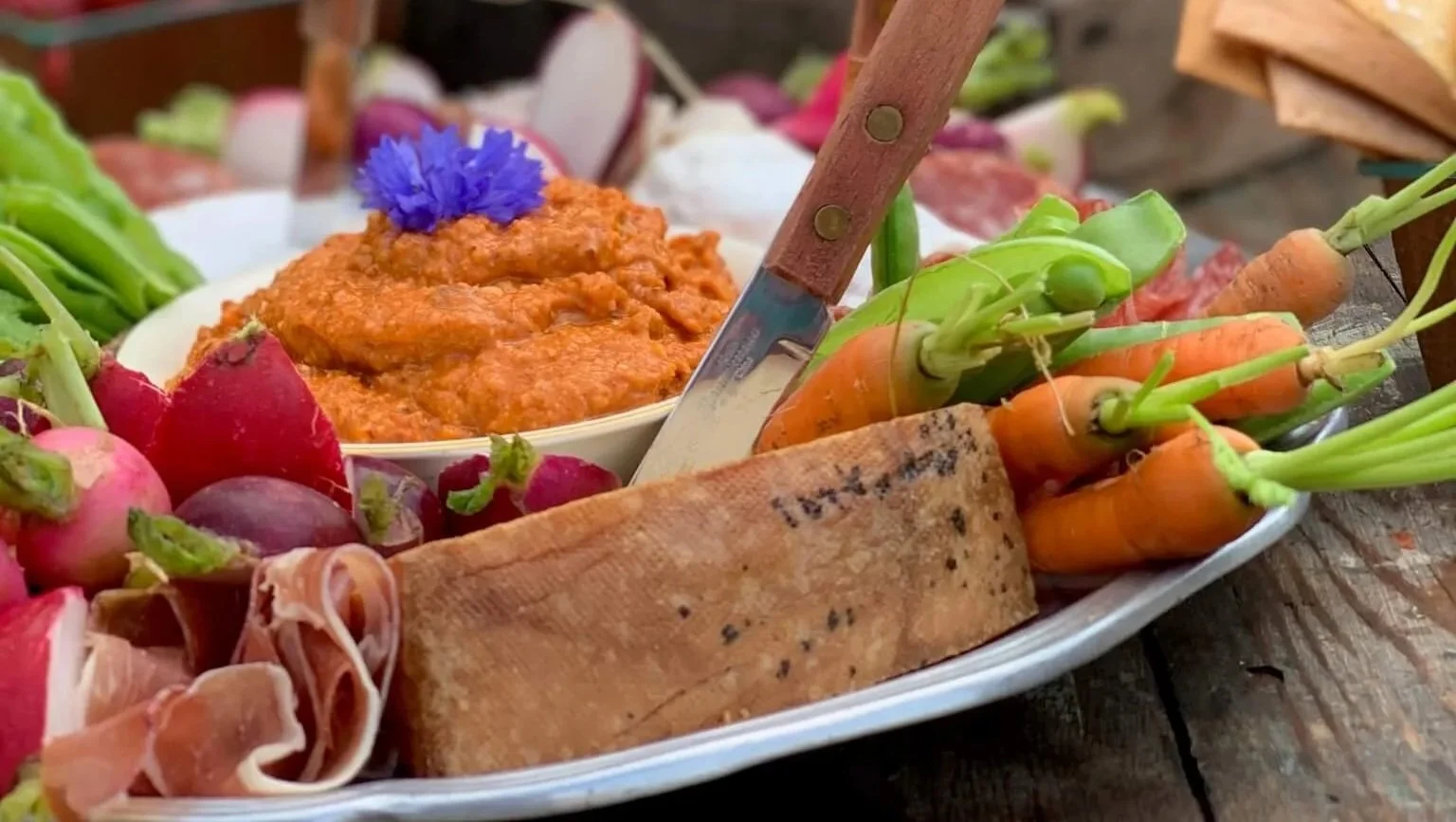 A serving platter with fresh radishes, carrots, cured meat, a slice of bread, and a bowl of tomato sauce garnished with an edible flower.
