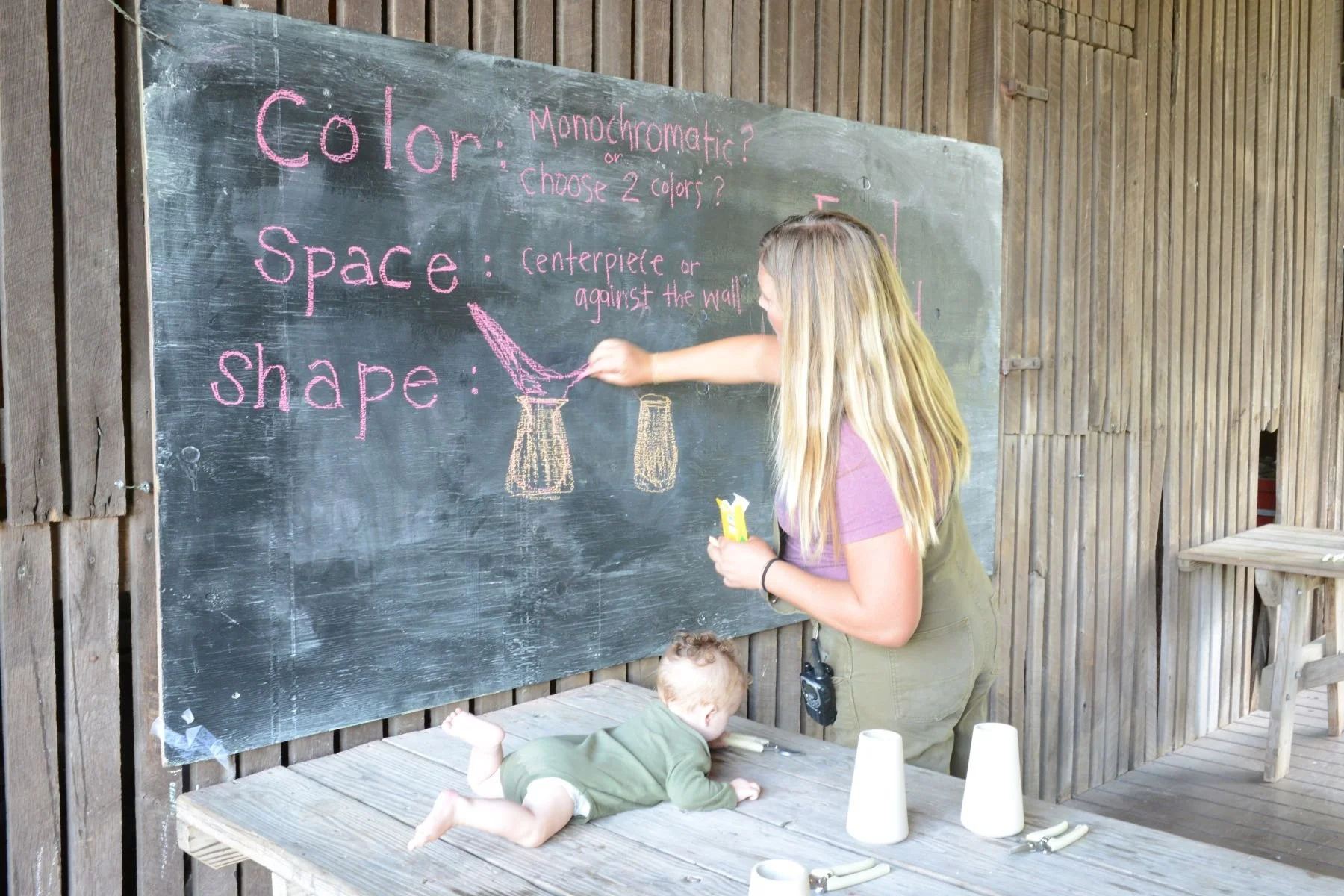 A woman and a child interacting near a chalkboard with colorful chalk drawings and writing about color, space, and shape. The woman is standing and drawing on the chalkboard, while the child is lying on a wooden platform below, reaching towards chalk