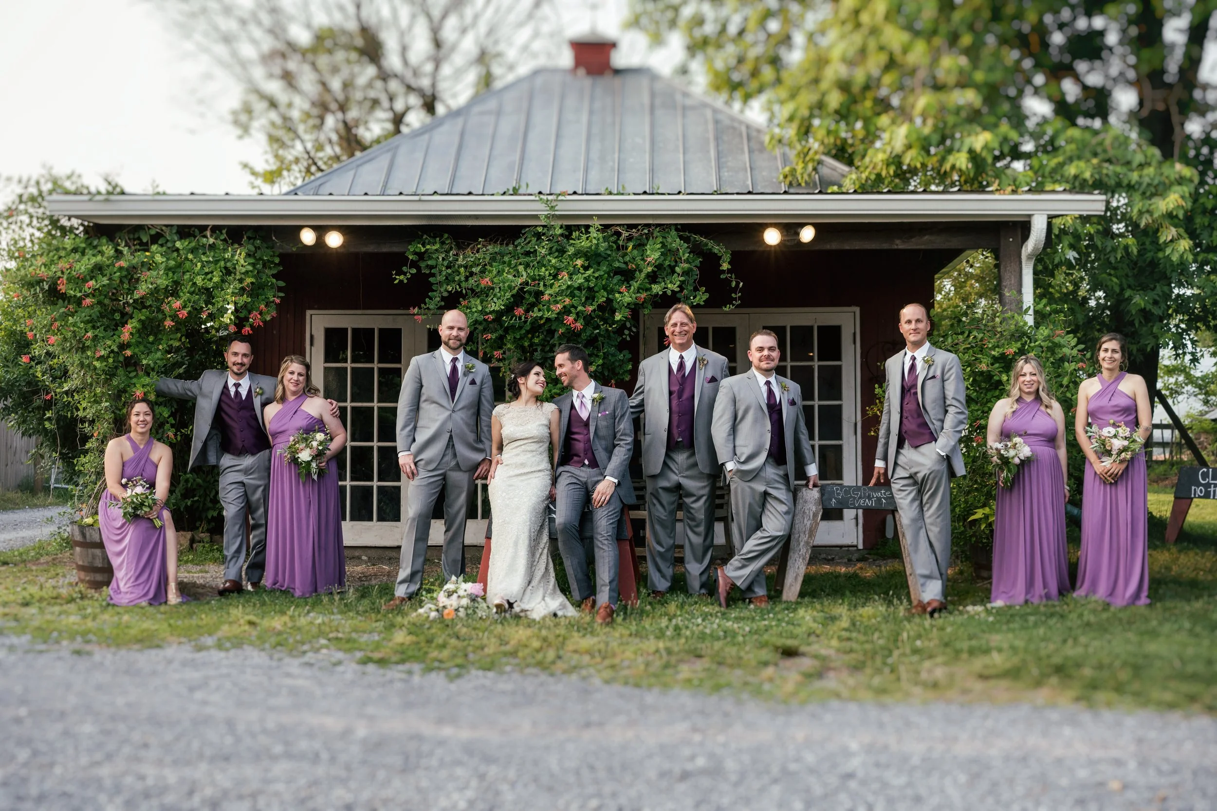 A group of people in formal attire standing outside a rustic barn, celebrating a wedding. The bride and groom are at the center, smiling and holding hands. Bridesmaids in purple dresses and groomsmen in gray suits with purple vests are around them, s