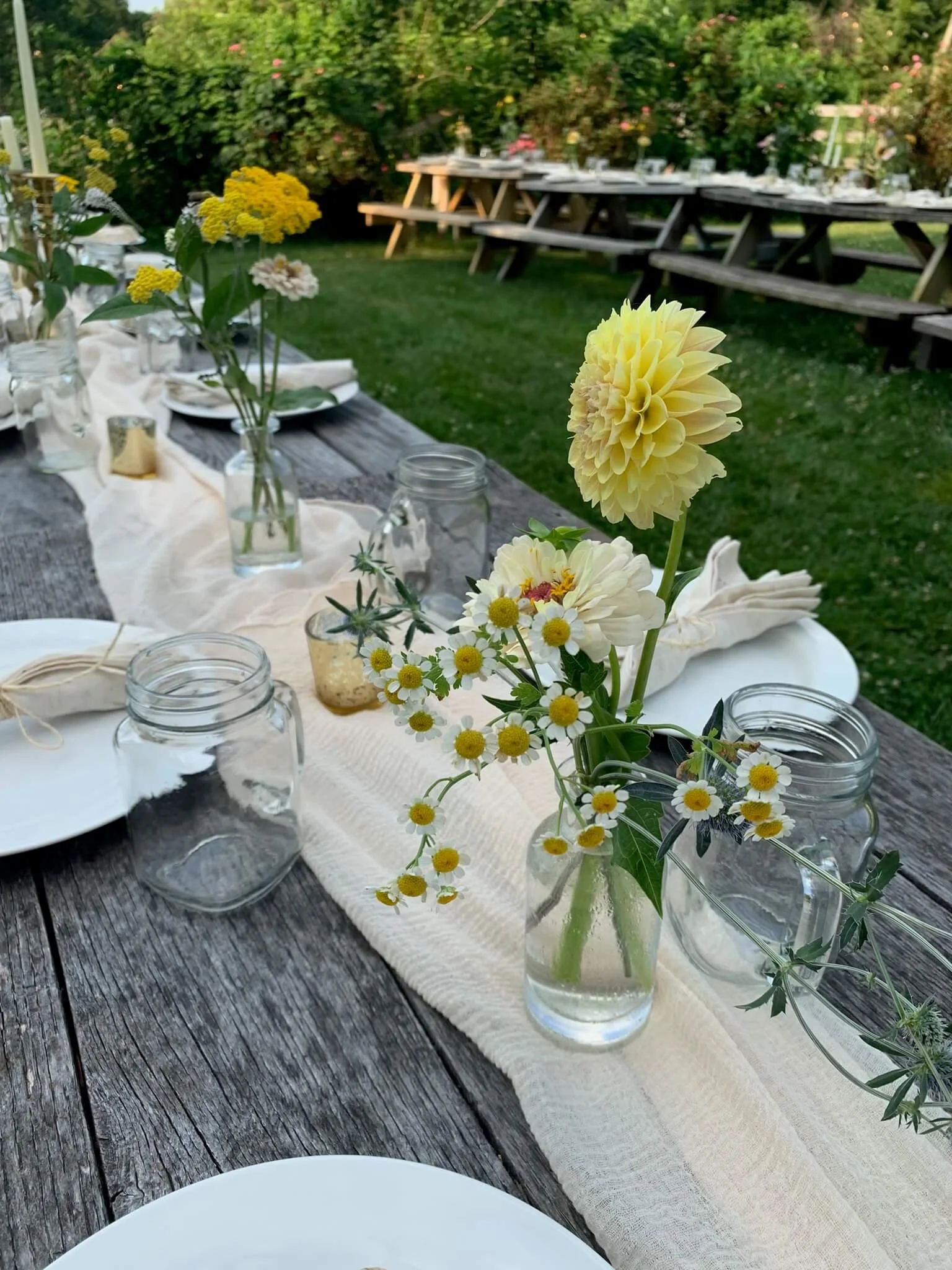 Outdoor table setting with flowers in glass jars on a rustic wooden table and picnic tables in the background, surrounded by greenery and blooming bushes.