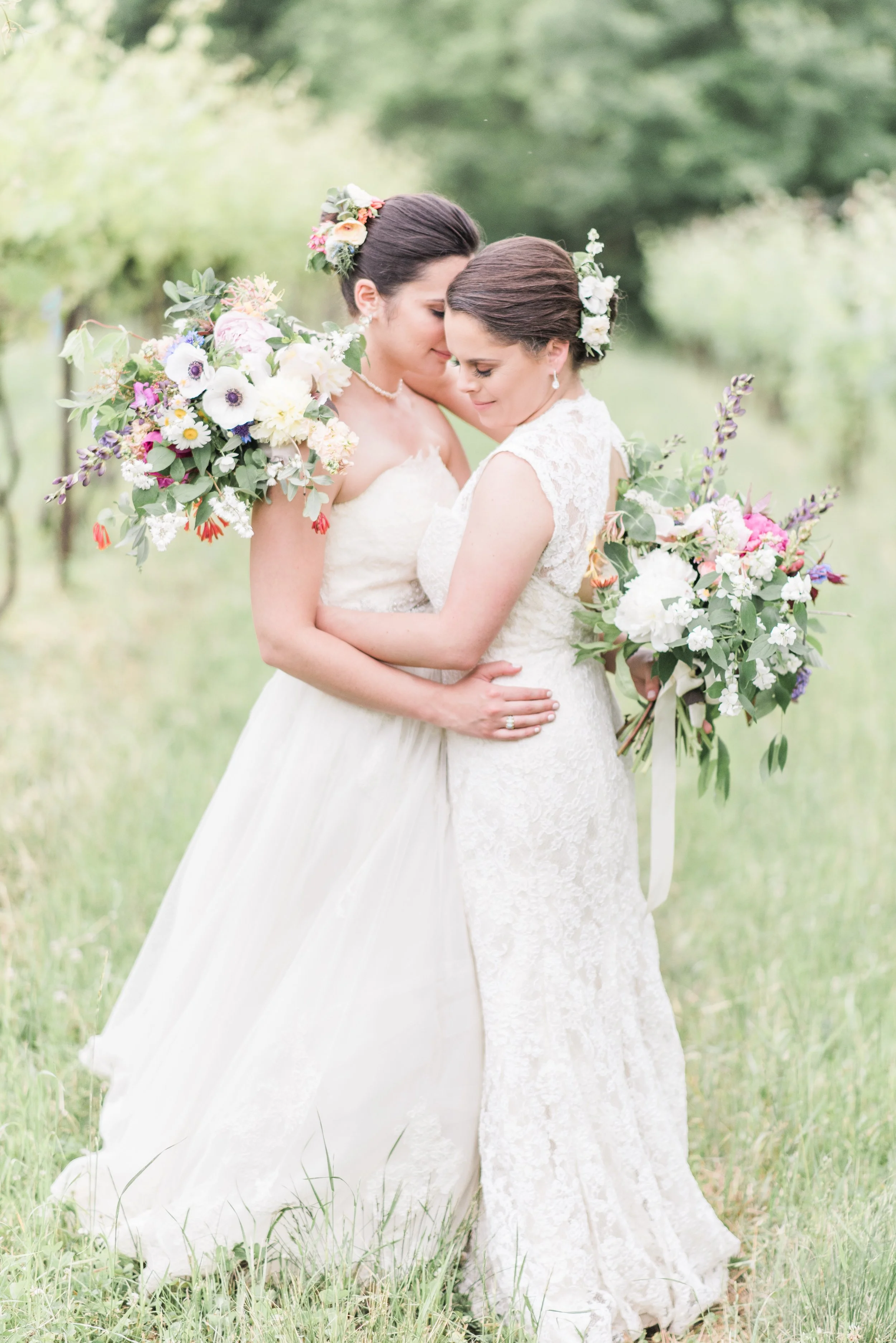 Two brides in wedding dresses standing close together outdoors, holding bouquets of flowers, with green trees and grass in the background.