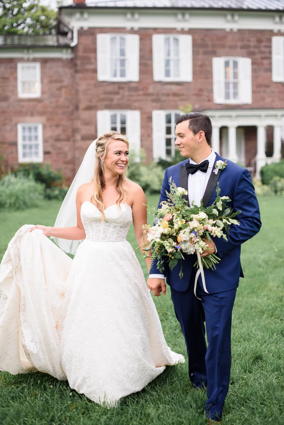 A bride and groom stand outdoors on a lawn, smiling at each other. The bride wears a strapless white wedding gown and veil, holding the train of her dress. The groom wears a navy blue tuxedo with a black bowtie, holding a large bouquet of flowers. A 