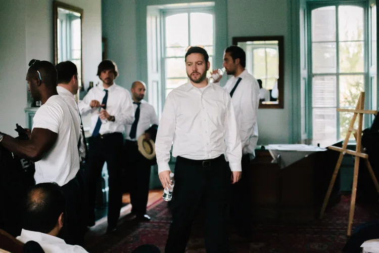 Group of men dressed in white shirts and black pants, some holding drinks, in a room with large windows and a vintage decor, appearing to be at a social or professional gathering.