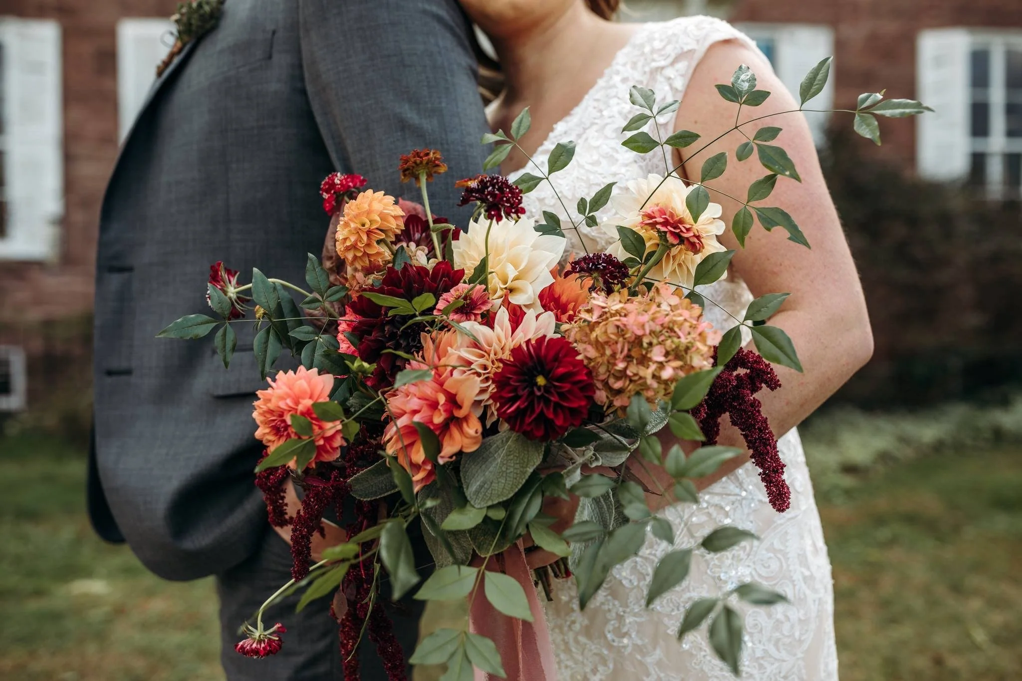 Close-up of a bride holding a colorful bouquet of flowers, standing next to a groom in a gray suit, with a brick building and windows blurred in the background.