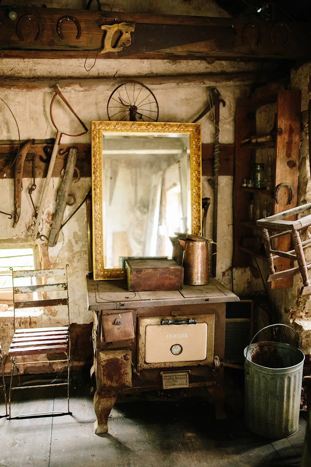 An antique rustic kitchen scene with a vintage wood stove, a large mirror with a gold ornate frame, old tools hanging on the wall, a small wooden shelf with jars, and a green trash can, all in a weathered, cozy setting.