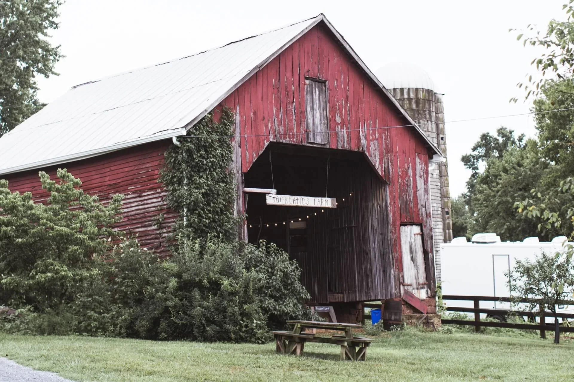 A weathered red barn with a metal roof, surrounded by green trees and bushes, with a small wooden picnic table in front and a sign reading 'Rocklands Farm' hanging near the entrance.