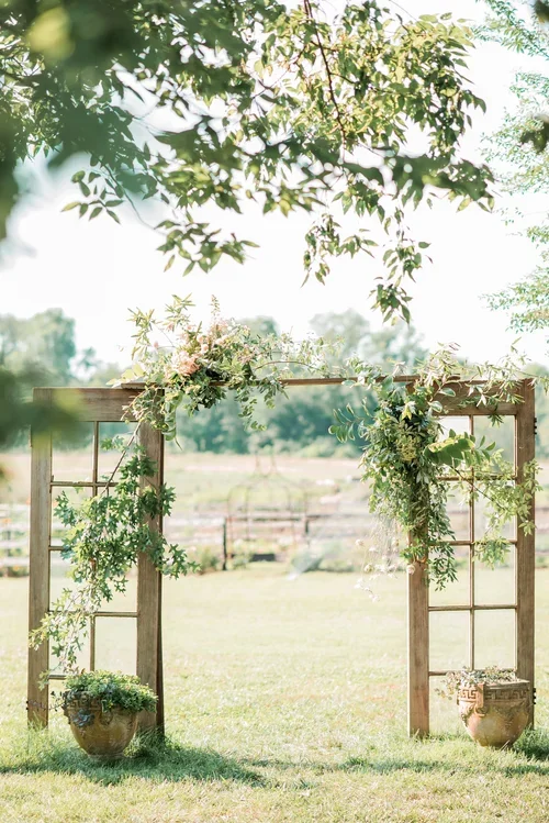 Wooden arch decorated with greenery and flowers, set outdoors in a grassy field.