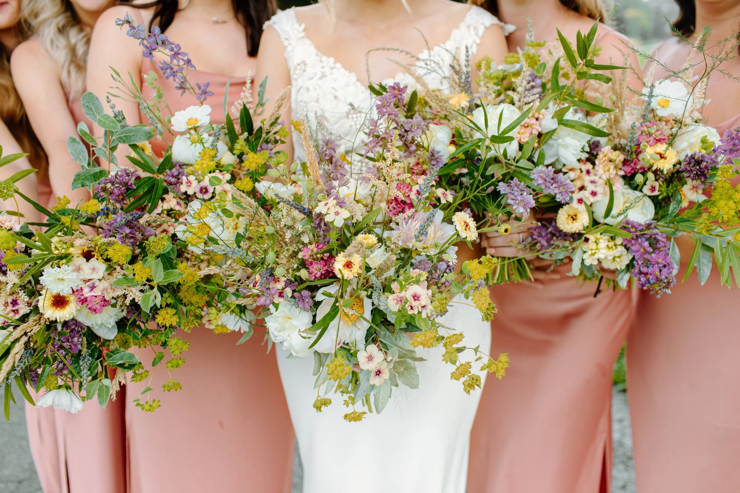 Bride holding a large bouquet of mixed colorful flowers, surrounded by women in pink dresses.