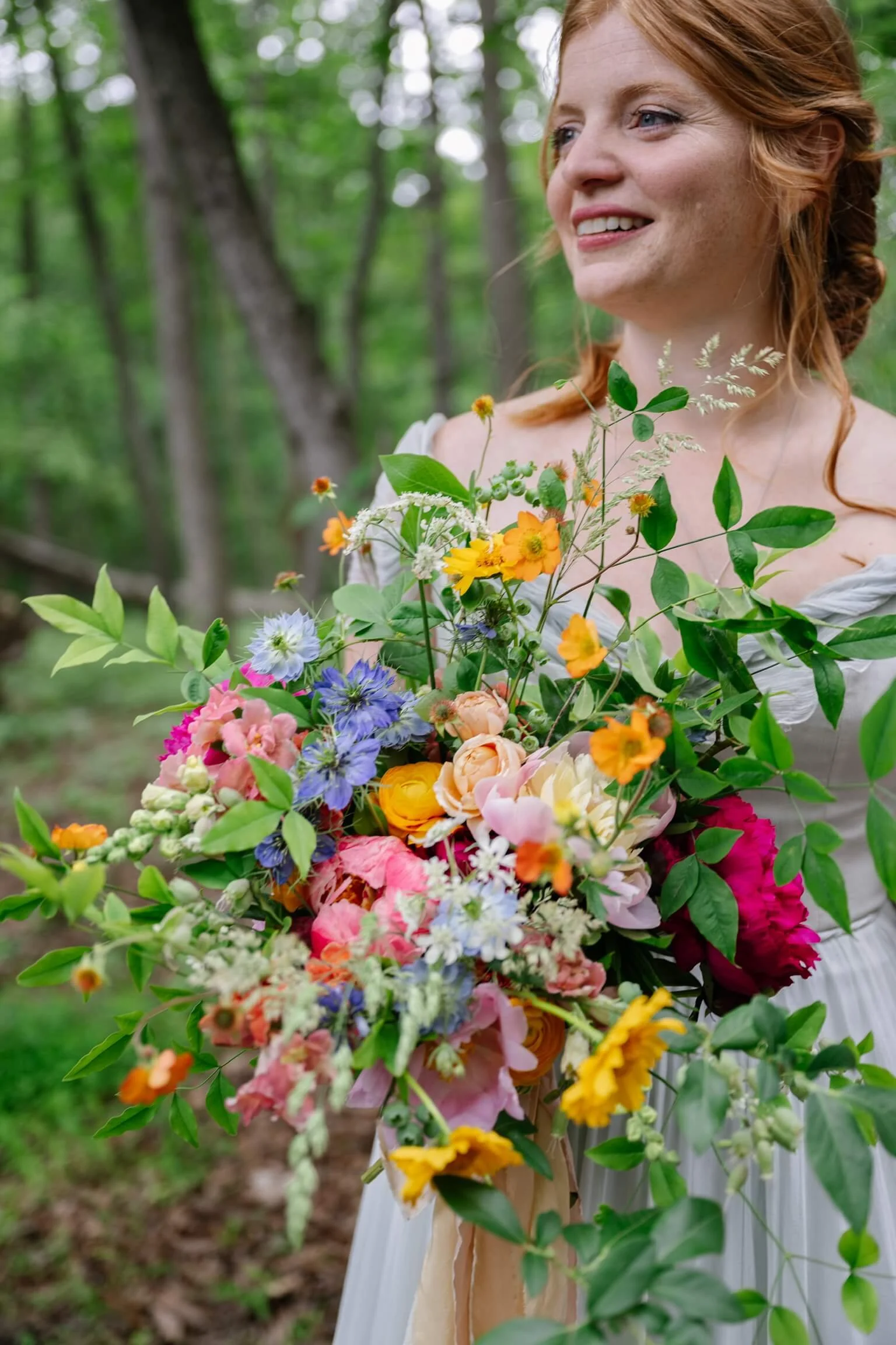 A woman with red hair and a white dress holding a large colorful bouquet of flowers outdoors in a forest setting during a wedding at Rocklands Farm Winery