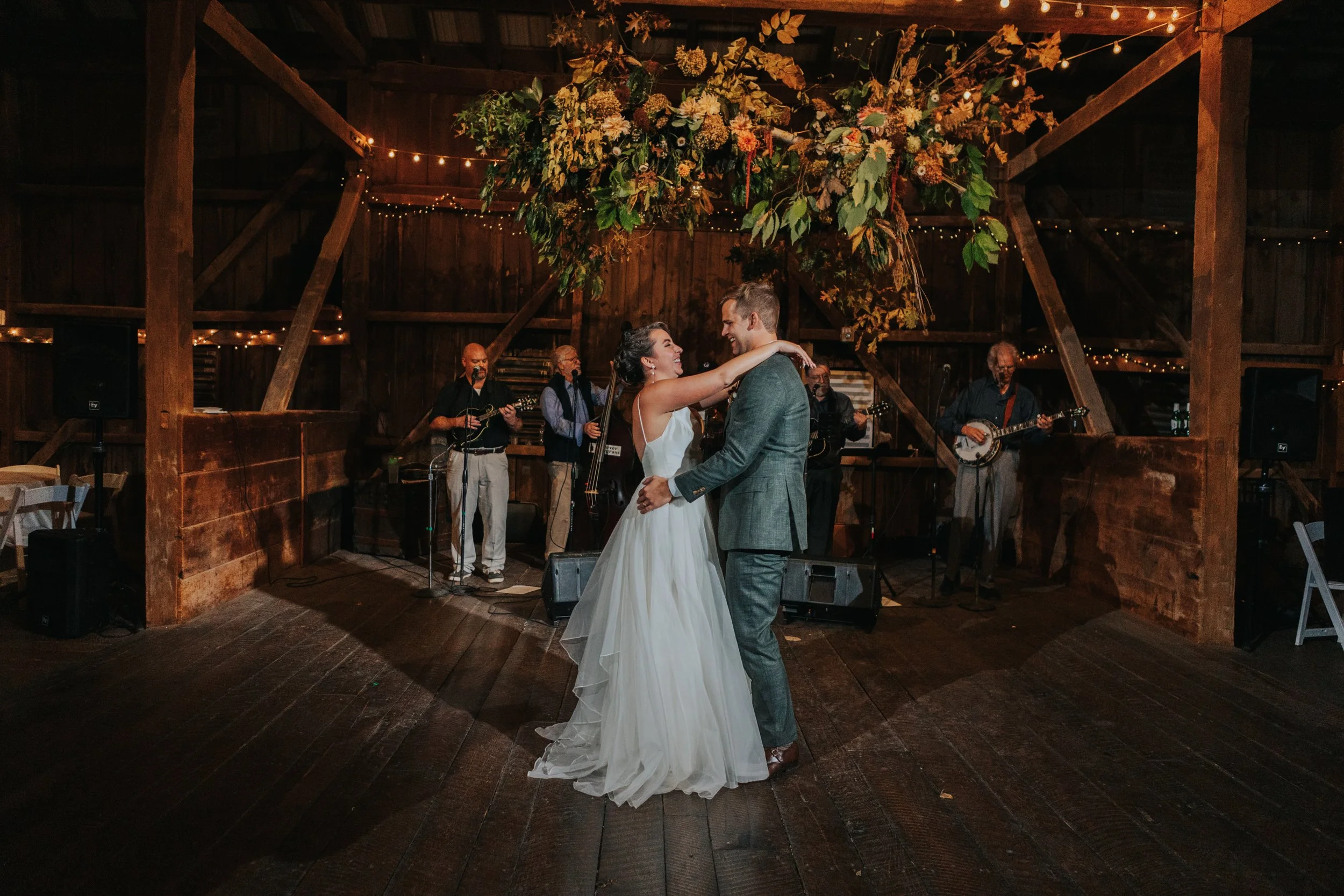 A bride and groom dancing at their wedding reception in a rustic barn decorated with floral arrangements and string lights, with a live band playing in the background.