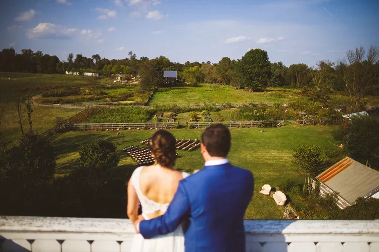 A couple dressed in wedding attire standing on a balcony overlooking a rural farm landscape with fields, trees, and a barn.
