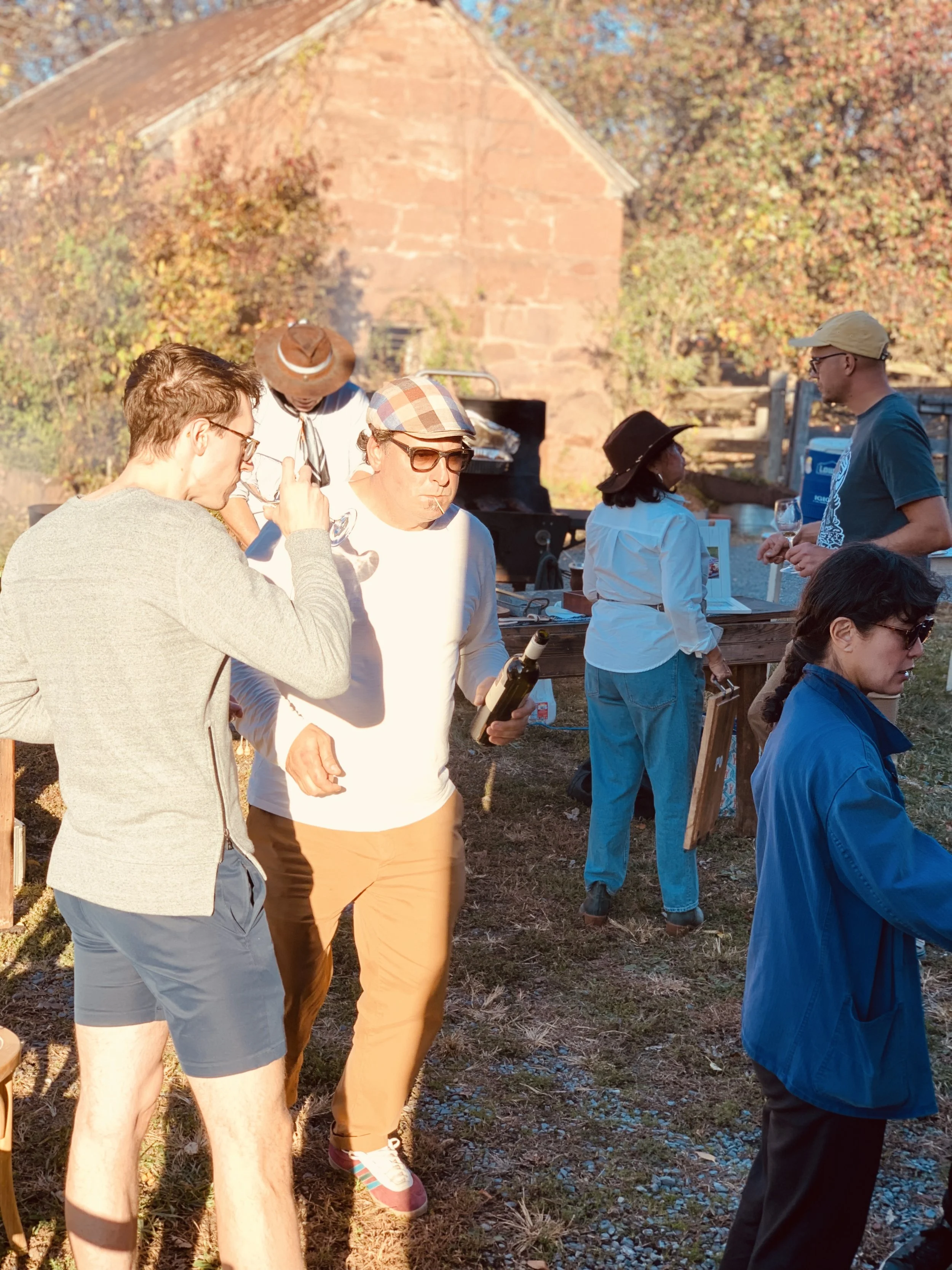 Group of people outdoors in warm sunlight, some drinking wine, with a rustic brick building and trees with autumn leaves in the background.