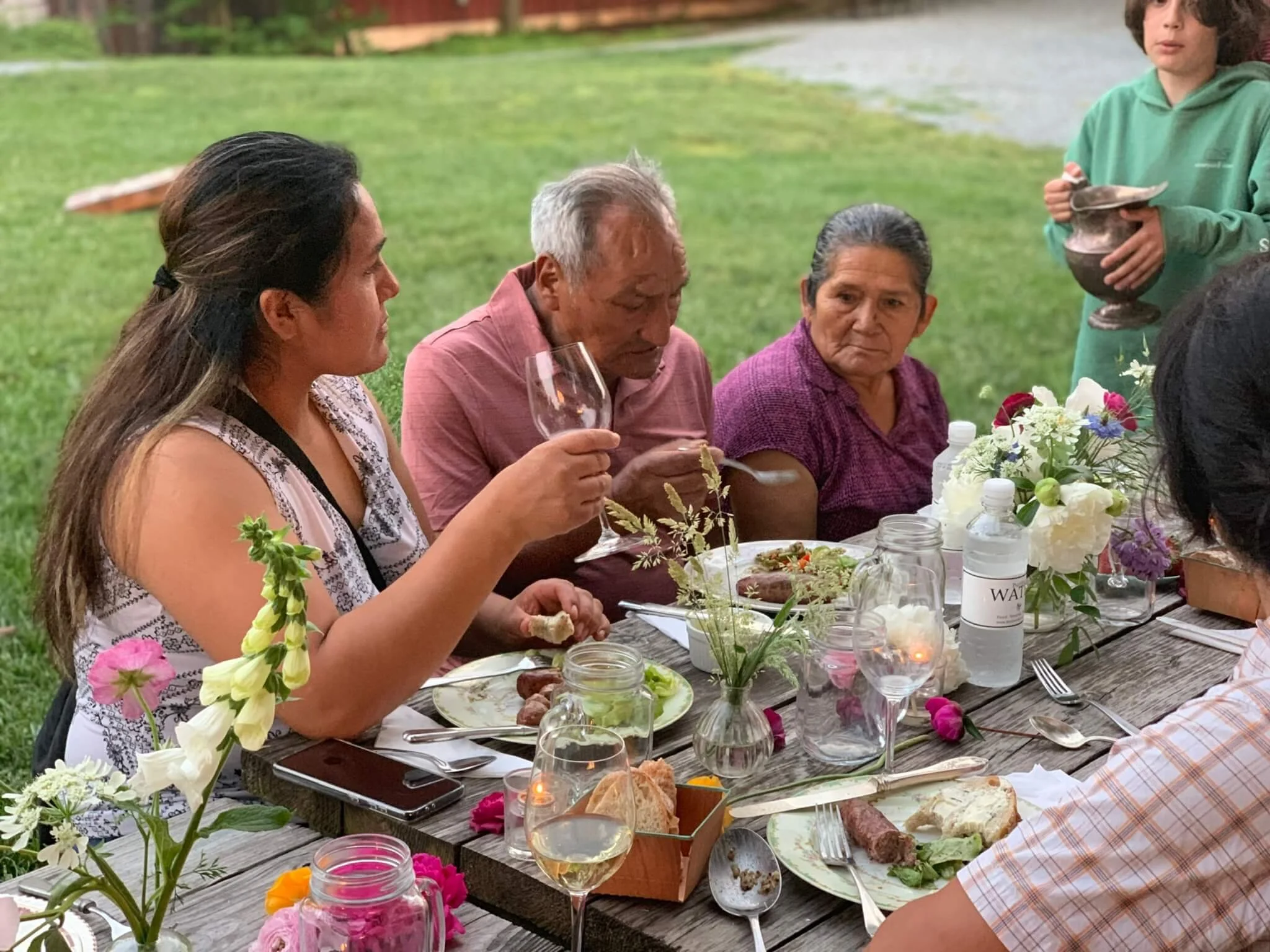 People sitting at a long outdoor table for a meal, with flowers, plates, and drinks, in a grassy area near a body of water.