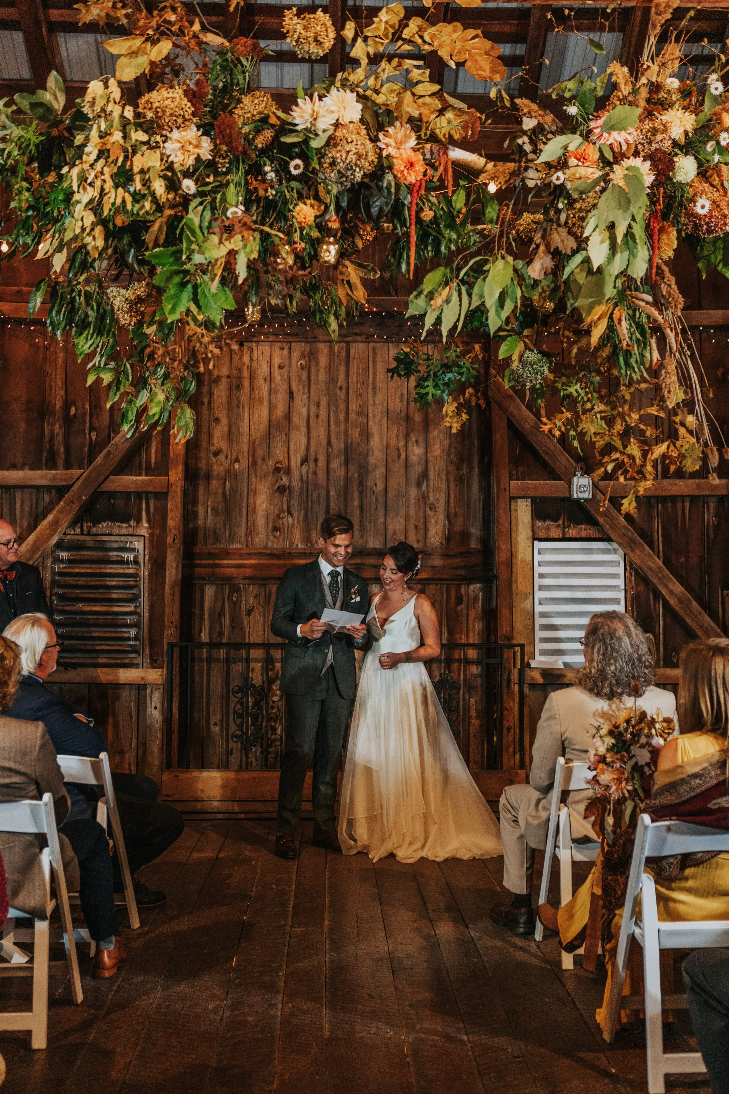 A wedding ceremony taking place in a rustic wooden venue, with the bride and groom standing at the altar under a large floral and leaf arrangement. Guests are seated in white chairs, watching the couple, and the decor features warm, autumnal tones.