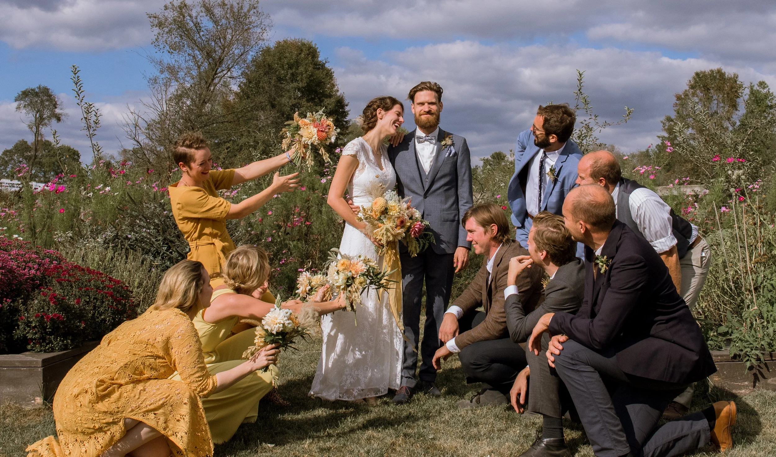 A wedding ceremony outdoors with a bride and groom smiling at each other, surrounded by friends and family. The bride holds a bouquet, and the group is engaged in a joyful moment in a garden setting.