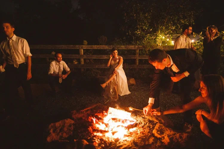 People gather around a campfire at night, some sitting and some standing, with trees and a wooden fence in the background.