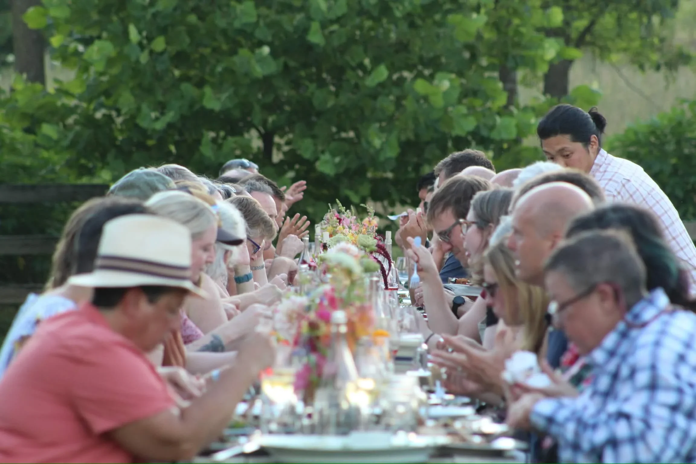 People sitting at a long outdoor table during a social gathering, with greenery in the background.