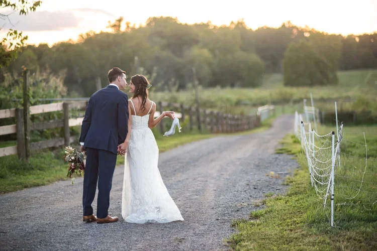 A newlywed couple holding hands and walking on a country road at sunset, with the bride in a white wedding dress and the groom in a dark suit, surrounded by greenery and a wooden fence.