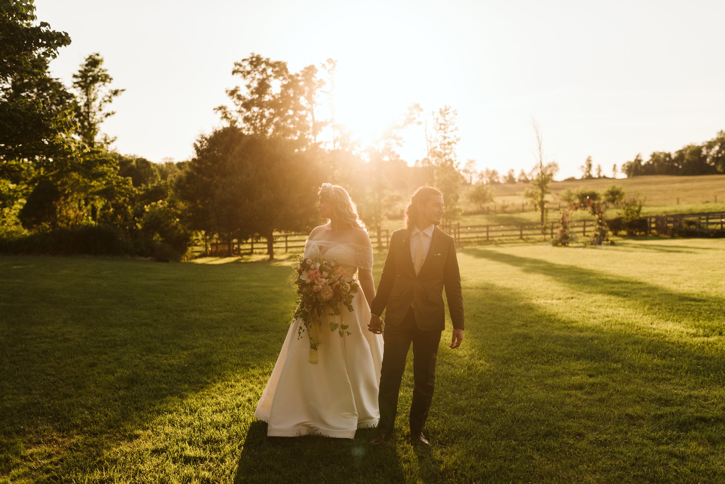 A bride and groom walking outdoors at sunset, holding hands; the bride is in a white wedding gown holding a bouquet, and the groom is in a dark suit, in a green field with trees and wooden fence in the background.