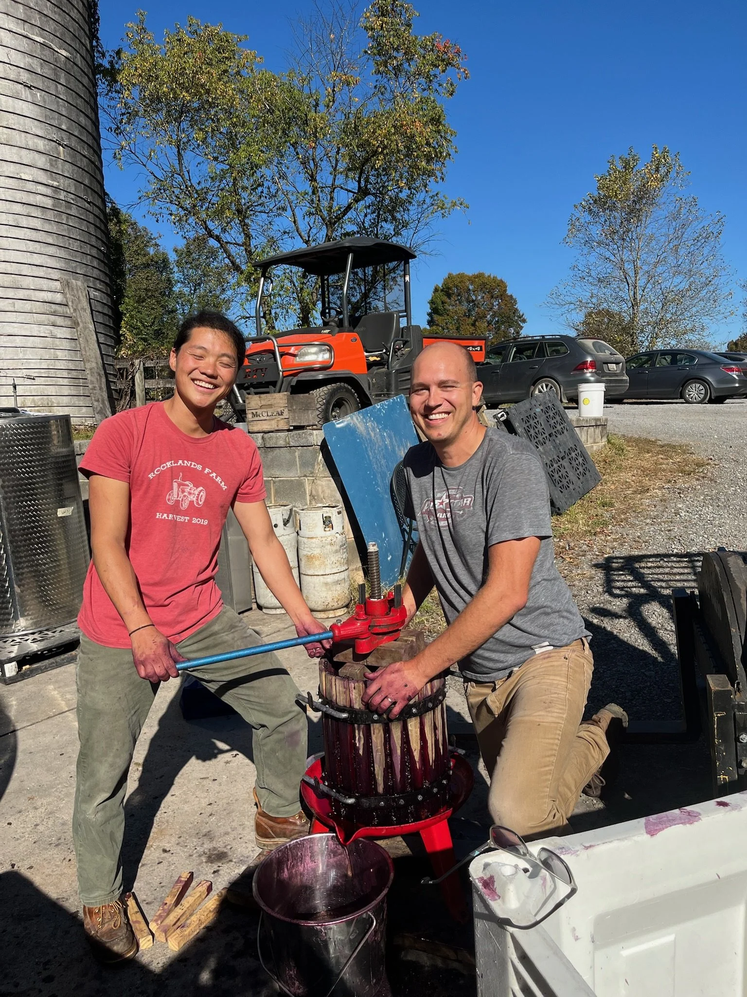 Two people, a woman and a man, smiling and working together outdoors on a cider press. The woman is wearing a red shirt and khaki pants, and the man is wearing a gray shirt and khaki pants. There are cars and trees in the background.