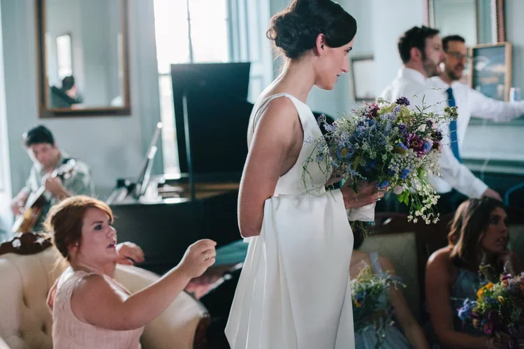 A woman in a white dress holding a bouquet of flowers at a social gathering or wedding reception, with other people engaging in conversations and playing music in the background.