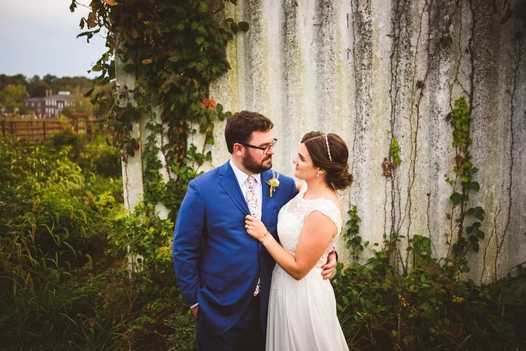 A bride and groom standing close together outdoors, in front of a weathered wall with vines, during their wedding, with greenery and trees in the background.