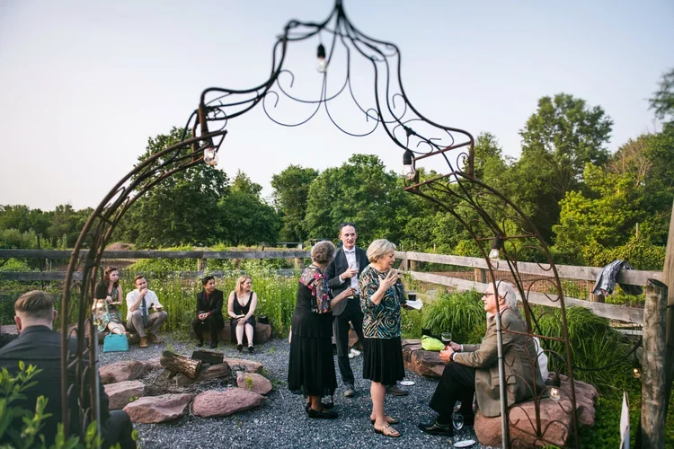Group of people gathering outdoors around a fire pit, some standing and some sitting, under a metal archway with string lights, with trees and a wooden fence in the background.